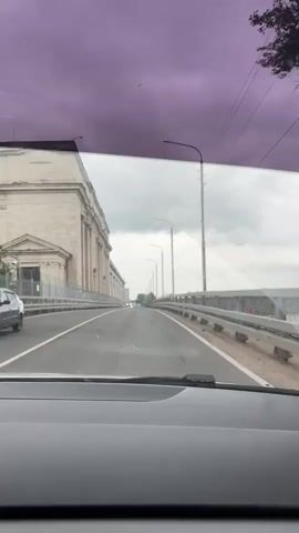 The video is a first-person perspective from inside a car driving across a bridge. The sky is overcast with purple-tinted clouds. To the left of the road, there is a large, old stone building with columns. The road itself is paved and has white lines marking the lanes. There are streetlights and power lines visible along the side of the road. In the distance, other cars can be seen on the bridge. To the right of the road, there is a railing with decorative metalwork, and beyond that, a body of water. The car's dashboard and side mirror are visible in the foreground.