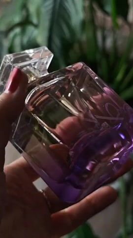 A hand with red nail polish holds a clear glass perfume bottle with a gradient of purple color at the bottom. The bottle has a faceted design and a clear cap. The background is blurred and shows green foliage.