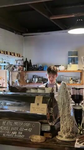The video shows a scene inside a small coffee shop. A young man with brown hair stands behind the counter, working with a coffee machine. He wears a light-colored shirt and an apron. Behind him, shelves are stocked with various items, including jars, bottles, teapots, cups, and kitchenware. A water dispenser is also visible on one of the shelves. In the foreground, a hand in a blue-striped shirt waves towards the barista. A sign on the counter reads "WE STILL PREFER CASH! PURCHASES UNDER RCO add R2 AMEXCARD ADD 3% ALL RIGHTS RESERVED". There's also a small sign indicating a charge for takeaway cups used for sitting. A decorative object resembling a white, petrified tree trunk stands on the counter next to the coffee grinder. A person's hand holding a straw is visible on the right. The overall lighting is dim, with a single lamp illuminating the counter area.