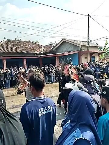 This is a video clip showing an outdoor event in Ngadirejo, Indonesia. A crowd of people is gathered, with many in the foreground looking towards a stage area covered by a large tent. The tent has a blue banner with yellow lettering that appears to say "GELAR BUDAYA & BERSIH QUSUN SID..." The banner is draped over the tent.

Behind the tent, a large speaker system on a metal truss is visible, indicating a performance or announcement is likely taking place. The weather appears to be sunny and bright, suggesting it is daytime. People are dressed casually. In the foreground, some individuals are wearing headscarves and traditional Indonesian clothing. The overall atmosphere seems to be one of community gathering and cultural celebration. The presence of a stage, sound system, and a banner suggests a planned event, possibly a cultural festival or a local gathering.