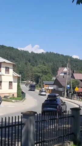 A black fence is in the foreground. A curved road goes uphill into a village. A white and tan building is on the left, and houses are on the right. A bus and several cars drive on the road. Trees and a mountain covered in trees are in the background. The sky is blue with some white clouds.