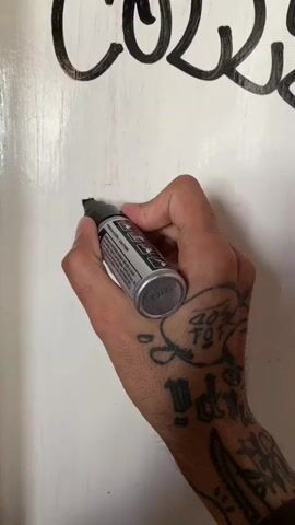 A close-up shot shows a hand with tattoos writing on a white surface with a black marker. The hand is holding the marker and moving it smoothly to form the word "MEMOIRS". Above this word, part of another word is visible, possibly "COLD". The background is plain white, suggesting a whiteboard or a clean surface. The hand appears to be the main focus, with the writing as the primary action.
