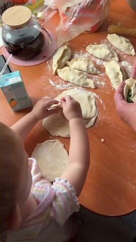 The video captures a close-up overhead view of a wooden table where two individuals, a child and an adult, are engaged in preparing dough. Flour is dusted across the surface, with several pre-made dumplings scattered on the right side of the frame. The child, facing away from the camera and wearing a striped shirt, uses both hands to manipulate a piece of dough, seemingly attempting to fold it. The adult's hands, positioned on the right side of the frame, are actively working with another piece of dough, demonstrating a folding motion. In the background, a glass teapot filled with dark liquid, a pink circular placemat, and a carton of orange juice are visible. The lighting suggests an indoor setting, likely a kitchen, with natural light entering from the side.