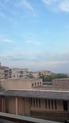 This media file captures an urban skyline against a pale blue sky with wispy clouds, suggesting a serene atmosphere. The primary subject is an airplane in flight, a small silhouette against the vast expanse of the sky. Below the aircraft, several buildings are visible, their architecture suggesting a European setting, possibly residential apartments with balconies and awnings. The time of day appears to be either early morning or late afternoon, given the soft, diffused light and the colors in the sky. The weather seems pleasant, with clear skies and only a few scattered clouds. The buildings are constructed with a light-colored facade, and some feature visible windows and balconies with striped awnings. The foreground shows the edge of a building with a dark, textured roof and vertical slats on its facade. The overall scene is one of quiet observation, perhaps from a window or balcony, looking out at the sky and the city. There are no people prominently featured in the video, and no visible text. The presence of an airplane in the sky could imply travel or the normal activity of an airport in the vicinity, but the scene itself is peaceful. The location context provided, Riudoms, Spain, further grounds the scene in a specific geographical area known for its Mediterranean climate and architecture, which aligns with the visual cues in the media.