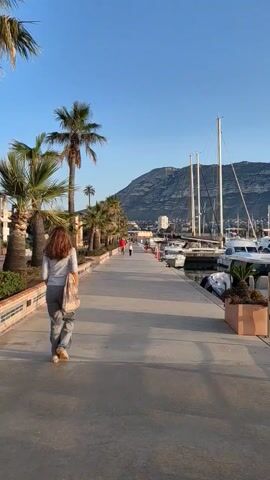 A woman walks away from the camera along a concrete pier on a sunny day.  She has long brown hair and wears gray pants and a white shirt.  She carries a large patterned bag over her shoulder. Palm trees line the left side of the pier, and moored boats fill the water to the right.  In the distance are several other pedestrians, buildings, and a large mountain against a clear blue sky.