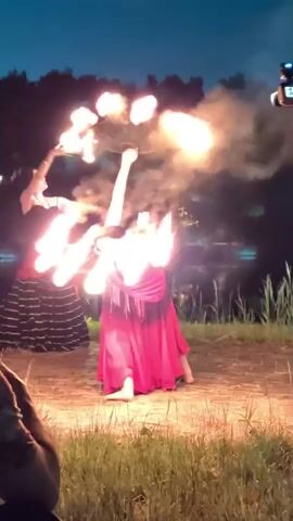 Two women are performing a fire dance outdoors at night. The woman in the foreground is wearing a red skirt and black top, and is holding a fire prop that resembles a fan with multiple flames. The woman in the background is wearing a black top and a long black and red striped skirt, and is holding two fire props, one in each hand. Both women appear to be in motion, with the woman in the foreground bending backwards as she spins her prop. Smoke is visible in the air around the flames. The background shows dark trees and a body of water, with a faint light source visible on the left. A person's arm and shoulder are visible in the lower left corner of the frame, suggesting they are observing the performance.