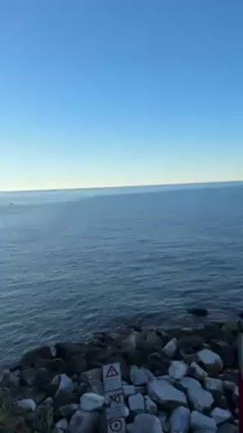 The camera is positioned above a rocky shoreline, offering a view of a calm ocean under a clear, blue sky. The horizon line is visible in the distance where the water meets the sky. A caution sign stands among the rocks in the foreground. The camera gradually pans to the right revealing more of the rocky terrain and ocean.