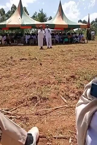A video shot from a low angle shows two adults in white standing in front of a seated crowd, likely at an outdoor event. The setting is a field with dry grass and soil, suggesting a rural area. Two large tents are set up in the background, providing shade for the seated people. The adults, dressed in white attire, appear to be speaking or gesturing to the audience. The crowd is diverse, with people wearing various clothing styles, although many seem to be dressed formally. The overall atmosphere seems to be celebratory or commemorative. The camera is relatively stable, providing a clear view of the scene.
