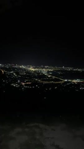 A nighttime aerial view of a sprawling city illuminated by countless lights. The lights form a dense, radiant carpet across the landscape, with brighter concentrations indicating major thoroughfares and commercial areas. The sky above is dark and clear, providing a stark contrast to the urban glow. The foreground is obscured by darkness, suggesting the vantage point is from a higher elevation, possibly a hilltop or mountain overlooking the city. The overall impression is one of vastness and vibrant nocturnal activity.