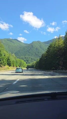 The video shows a view from the inside of a car driving on a highway. The road is paved with white lane markings and cars can be seen driving ahead. The surrounding environment is mountainous and covered with dense green trees. The sky is blue with scattered white clouds. The sun is shining, casting shadows on the road.