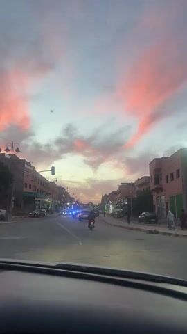 The view from the dashboard of a vehicle driving down a city street shows a row of brick buildings, with various storefronts. Cars are parked on both sides of the street, and vehicles, including a motorcycle, are traveling in both directions. A pedestrian walks along the sidewalk on the right. A traffic light can be seen. The sky is a mix of blues and grays, with patches of pink and orange in the clouds at dusk. There are lampposts lining the street.