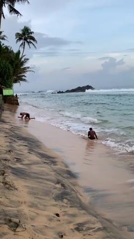 The video shows a sandy beach with two men at the shoreline. One man kneels on the sand, then leans forward and touches the ground with his head. The other man sits in the water at the edge of the beach. Palm trees and greenery are visible to the left of the frame. In the ocean, waves are breaking, and there are rock formations further out. The sky is overcast with clouds.
