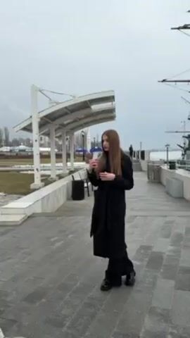 The video shows a young woman standing on a paved walkway next to a large ship replica. She is wearing a long black coat. The ship is docked behind a black metal fence, and it has multiple masts and intricate decorations. The sky is overcast, and there are other people walking in the background. The woman appears to be recording a video, possibly for social media.