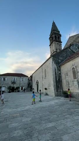 This video captures a serene scene in Perast, Montenegro, likely during the late afternoon or early evening as suggested by the soft, diffused light and clear blue sky with scattered clouds. The primary subjects are three young boys playing in a spacious, stone-paved square.

In the foreground, two boys are engaged in play. One boy, wearing a blue shirt and yellow shorts, appears to be kicking a ball or practicing some movement with his feet. Another boy, in a green shirt and shorts, stands nearby. A third boy, in a lime green shirt and red shorts, stands further to the right, observing or perhaps waiting his turn. Their presence injects a sense of lively childhood activity into the otherwise tranquil setting.

Dominating the background is a magnificent, ancient-looking church with a prominent bell tower. The church is constructed from weathered stone blocks, showcasing a rustic architectural style. Its tall, pointed bell tower, capped with a spire, reaches towards the sky, a stoic sentinel over the town. The soft sunlight highlights the textures of the stone and the details of the church's facade, including arched windows and decorative elements.

To the left of the church, a building with red-tiled roofs and traditional architecture is visible, hinting at the historical charm of Perast. The square itself is vast and paved with large, irregular stone slabs, creating a timeless ambiance. Potted plants are strategically placed, adding touches of greenery to the scene.

The overall mood is peaceful and idyllic, with the children's play adding a touch of youthful energy. There is no visible text within the frame. The video effectively conveys the historic atmosphere of Perast and the simple joy of children at play in a beautiful, timeless setting.