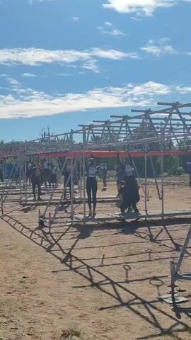 The video shows an outdoor obstacle course on a sunny day with a blue sky and scattered clouds.  A group of people are navigating a horizontal monkey bar-like obstacle constructed from metal scaffolding. Some individuals are hanging from the bars, while others are in mid-movement, either swinging or preparing to move to the next bar.  The ground is a dry, sandy dirt.  Shadows from the scaffolding structure are cast across the ground.  The people are wearing athletic clothing.