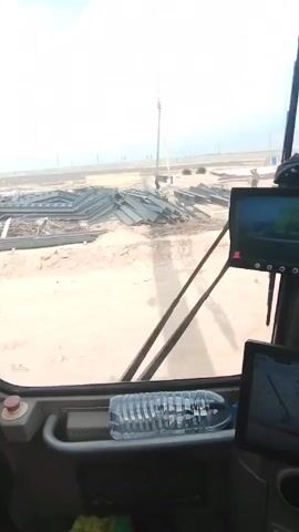 Here's a description of the video:

The video shows a construction site, as viewed from inside the cab of a construction vehicle (likely a crane or similar heavy equipment). Outside the vehicle, there are stacks of metal beams and a worker visible. The camera pans to the right, revealing partially constructed metal framework for a building. Workers are present on the construction site. Signs with warnings in Chinese, Russian, and English are visible inside the cab. The ground is mostly sand and dirt. The weather appears to be overcast or hazy.