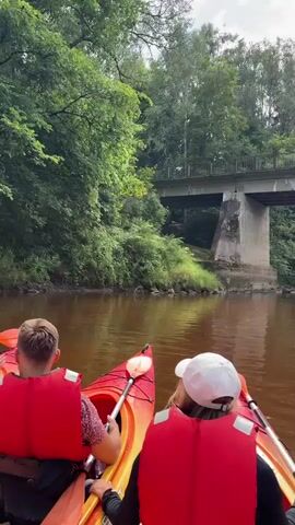 Two people in kayaks paddle down a muddy river. They are wearing red life vests. The person in the foreground is wearing a white cap. The kayaks are orange and yellow. A concrete bridge with a metal railing spans the river in the background. Dense green trees line the banks of the river. The sky is cloudy.