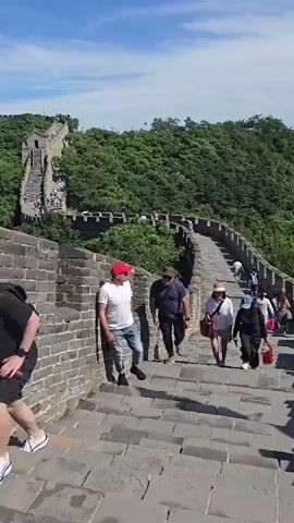 A group of tourists are walking up a steep stone staircase on the Great Wall of China. The wall is made of grey stone bricks and stretches into the distance, surrounded by lush green trees. The sky is a bright blue with a few white clouds. The people are dressed in casual clothing, and some are carrying bags. The camera is positioned behind the group, looking down the stairs.