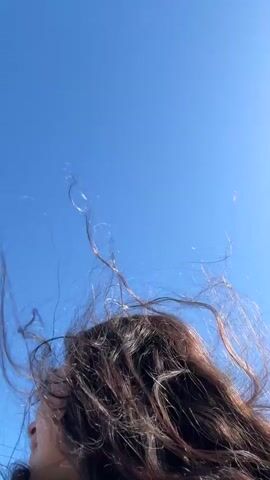 A young woman with long, wavy, dark hair is standing on a sidewalk by a street, speaking into the camera. Her hair is being tossed by the wind. A car drives by behind her. The sky is blue. She is smiling slightly at the end of the video.
