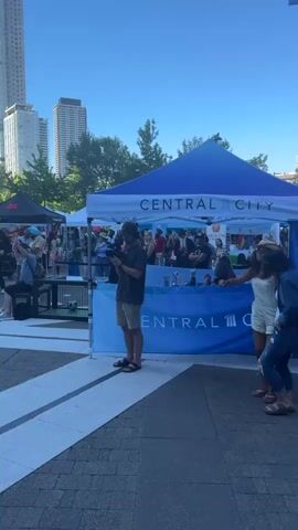 A daytime outdoor event with a blue tent displaying "CENTRAL CITY" branding. Two women are dancing near the tent. The woman on the left wears a straw cowboy hat, overalls, and flip-flops. The woman on the right wears a denim jacket, ripped jeans, and sandals. Several other people are visible in the background, some standing behind a table inside the tent. The ground is paved with bricks and white lines. The sky is clear and blue, with buildings and trees visible in the distance.