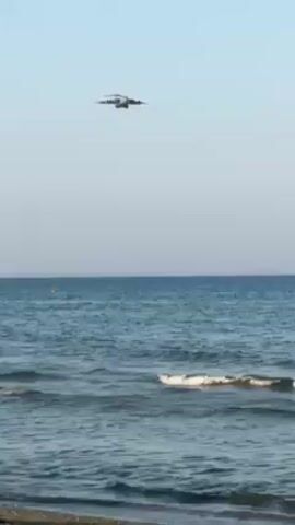 Certainly! Here's a detailed description of the video:

**Scene:**

The video shows a beach scene with a large cargo plane approaching from over the sea.

**Environment:**

*   **Sky:** The sky is clear with a gradient of blue, lighter towards the horizon and deeper blue overhead.
*   **Sea:** The sea is calm, with small waves breaking near the shore. The color is a deep blue-green.
*   **Beach:** A sandy beach is visible in the foreground.

**Objects:**

*   **Cargo Plane:** A large, military-style cargo plane is the main subject. It's flying relatively low over the water.
*   **Buoy/Marker:** A small yellow buoy or marker is visible floating in the sea.
*   **People:** There are people on the beach, though they are difficult to make out in detail.

**Actions:**

*   The cargo plane is flying towards the viewer, approaching the shore.
*   Waves are gently breaking on the beach.

**Interactions:**

*   There may be people on the beach observing the plane as it approaches, but their interactions are not clearly visible.

**Overall Impression:**

The video captures the sight of a large plane flying low over the sea, creating a striking contrast against the calm water and blue sky.