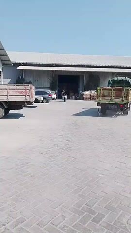 A person is riding a blue and white scooter from the center of the frame towards the viewer. The person is wearing dark clothing and a helmet. In the background, there is a large warehouse building with an open entrance. To the left of the warehouse, there is a red truck. To the right of the warehouse, there is a green truck loaded with what appears to be produce covered by a tarp. Several cars are parked near the warehouse entrance. The ground is paved with interlocking bricks. The sky is clear and blue.