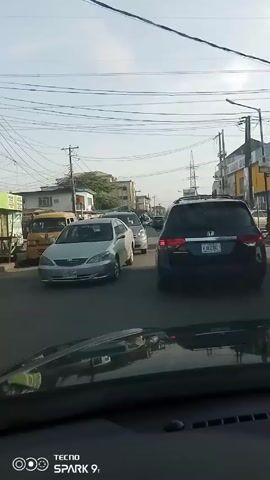 The view from inside a car shows city street traffic. There are multiple cars in front of the car and people visible on the sidewalks. A motorcycle drives by on the left. The sky is pale blue. Utility poles with wires run overhead. Buildings of varying heights are seen in the background. A sign for “AZREZ” is visible on a building.