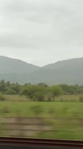 The video shows a blurred view from a moving train. The foreground features train tracks, and beyond them, a landscape of green fields and trees. In the distance, hazy mountains are visible under a cloudy sky. The overall impression is of a scenic journey through a rural or natural environment.