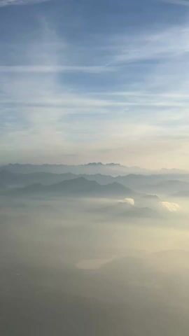The video shows an aerial view of mountains partially obscured by a layer of fog or haze, under a blue sky with thin, wispy clouds. The mountains are silhouetted in the distance, appearing as a series of ridges fading into the horizon. A lake or body of water is visible through a break in the fog.