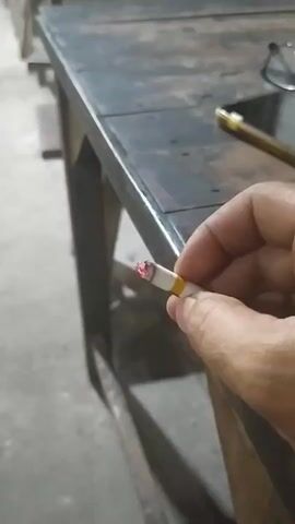 A close-up shot shows a person's hand holding a lit cigarette. The cigarette has a glowing red tip, indicating it is burning. The hand is positioned near the edge of a dark, wooden table. In the background, a pair of glasses rests on the table. The overall environment appears to be an indoor space, possibly a workshop or an office, with a concrete floor visible. The video is short, capturing only a few seconds of the scene.