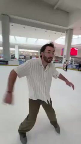 A person with dark hair and a mustache is ice skating on an indoor rink. The skater is wearing a beige, short-sleeved, striped shirt and brown pants. The person is holding their arms out for balance. The rink is surrounded by advertising panels and is located in what appears to be a large indoor shopping mall. Other people are on the ice in the background. The floor is white and the roof is white and high. Large white pillars support the roof.