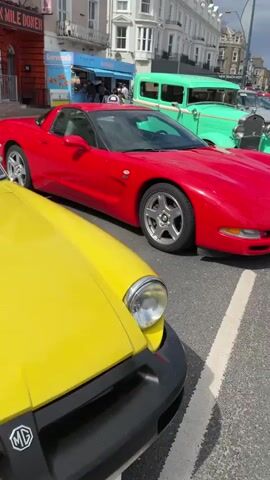 The video shows a street scene with a line of cars. A red Chevrolet Corvette is parked next to a yellow vehicle. Behind the Corvette are two vintage cars, one green and one silver. A young person takes a picture of the Corvette with a smartphone. People walk by, and a black-and-white police car is also visible in the distance. The scene is outdoors on a sunny day with some clouds. Buildings and a boardwalk are visible in the background.