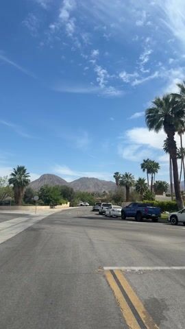The video shows a street scene on a sunny day. Several cars are parked along the side of the road. In the distance, there are mountains under a bright blue sky with scattered white clouds. Palm trees and other greenery are interspersed among houses and buildings along the street.