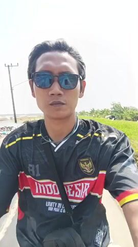 The video shows a person from the chest up, wearing sunglasses and a black jersey with red and yellow accents. The jersey has the word "INDONESIA" written across it and what appears to be a national emblem on the chest. The person is outdoors, with a bright sky in the background. To the left, there are some trees and a utility pole. To the right, there is greenery and vegetation. The person is looking directly at the camera. The video appears to be shot from a moving perspective, likely from a bicycle or motorcycle.