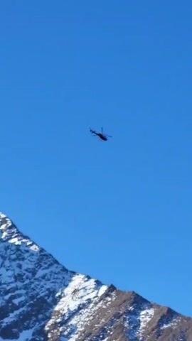 A helicopter flies across a clear blue sky, moving from left to right in the frame. The camera then pans downwards, revealing a majestic landscape of snow-capped mountains. The mountains are rugged, with dark rock faces partially covered in bright white snow, contrasting against the vibrant blue sky. The scene is set on a clear, sunny day, highlighting the stark beauty of the mountainous terrain. The location is Tengboche, Nepal. There is no visible text.