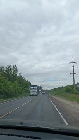A dashboard-mounted camera captures a forward-facing view of a road trip in Тополиный, Russia. The scene unfolds under a cloudy, overcast sky, suggesting a temperate climate, possibly late spring or summer, with daylight clearly present. The road, paved and two-laned, is bordered by lush green trees and dense foliage on either side, indicative of a rural or semi-rural setting.

Several vehicles are visible on the road. In the foreground and to the left, a white truck with a blue cargo container is traveling in the same direction as the camera. Further down the road, a white passenger car is also moving forward. As the video progresses, other vehicles appear and disappear from view, including a black car that briefly comes into frame. Power lines and poles are visible on the right side of the road, suggesting the presence of infrastructure.

The camera is positioned inside a vehicle, with the top portion of the dashboard visible at the bottom of the frame. This perspective provides a dynamic sense of movement as the vehicle navigates the road. The overall mood of the video is one of ordinary travel, capturing a moment of transit through a natural landscape. No text is visible in the footage.