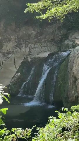 The video shows a small waterfall cascading into a pool of water. The waterfall is surrounded by rocks and lush green vegetation. The water appears clear and the scene is peaceful and serene. The sunlight filters through the leaves of trees, creating dappled patterns on the water and rocks.