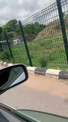 From a car's perspective, driving on a paved road with other cars ahead in line. A green wire fence runs alongside the road, separating the road from a grassy hillside. The sky is partly cloudy.
