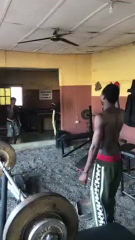 Two shirtless men are working out in a gym with old equipment. One man with green sweatpants is in the foreground doing stretches and the other is doing shadowboxing in front of the mirror. The gym looks old and is very basic.  There is a bench, a barbell on a stand, and a weight-stack machine in the background. There is also a sign that says “YOUR GYM TICKET IS HERE” in the background. There is a ceiling fan on.