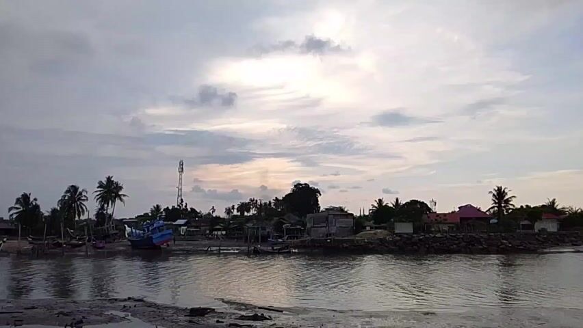 The video shows a coastal scene with several fishing boats docked or beached in a muddy area. The water is calm with reflections of the sky. The sky is overcast with clouds, with some light peeking through. Palm trees and other vegetation are visible along the shoreline, along with a few simple structures and a telecommunications tower. The camera seems to be on a vehicle driving along the coastline. The sound of the vehicle is clearly audible.