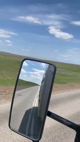 The video shows a road trip through a grassy landscape. The view is from the side mirror of a moving vehicle, showing the asphalt road and the passing landscape. The landscape consists of green fields under a blue sky with scattered white clouds. A small lake or pond is visible in the distance. The road has a guardrail alongside it. The video ends with the side mirror disappearing from the frame.