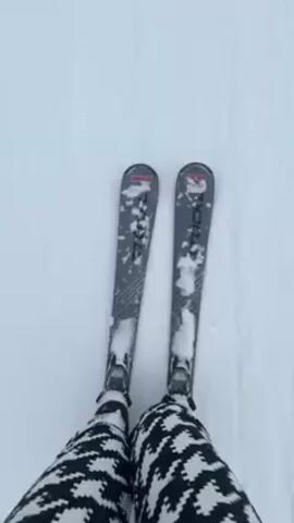 The video shows a first-person view of someone skiing down a snow-covered slope. The skier is wearing black and white patterned pants and grey skis. The snow is white and somewhat tracked. As the skier approaches the bottom of the slope, a building and other skiers become visible. The surrounding environment includes snow-covered hills and trees. Snow is falling.