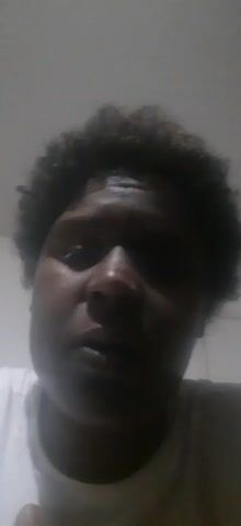 A close-up, dimly lit video shows a person with dark skin and short, curly dark hair. The individual is wearing a white t-shirt and looking directly into the camera with a serious expression. The background appears to be a plain, light-colored wall.