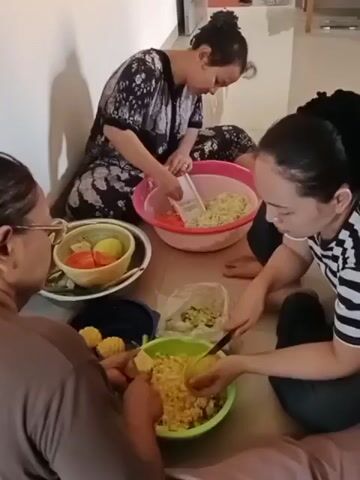 Three individuals are seated on the floor, preparing food. The person on the left is wearing glasses and a brown top, facing away from the camera. They are holding a yellow object, possibly corn on the cob. In front of them is a green bowl filled with yellow kernels, likely corn, and a dark blue container. The person in the middle is wearing a dark floral-patterned top and is seated facing right. They are using a grater to shred cabbage into a large pink bowl that also contains shredded carrots and possibly other vegetables. The person on the right is wearing a black and white striped top and is facing left. They are using a knife to cut a yellow object, appearing to be corn on the cob, into a green bowl containing corn kernels. There is a white plastic bag to their left, also containing shredded vegetables. A white bowl containing sliced tomatoes and yellow slices, possibly potatoes or more corn, is located between the person on the left and the person in the middle. The environment appears to be indoors, with a light-colored wall in the background and a shadow cast on it. The floor is light brown. The overall impression is of a casual food preparation session.