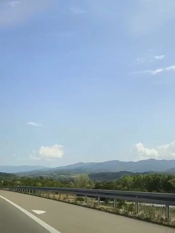 The video shows a view from a moving vehicle on a highway. The road curves to the left, with a white line marking the lane. A metal guardrail runs along the right side of the road. In the distance, there are rolling hills covered in green vegetation. The sky is a clear blue with a few scattered white clouds. Several vehicles are visible on the highway in the distance, including trucks and cars. The overall impression is of a scenic drive on a sunny day.