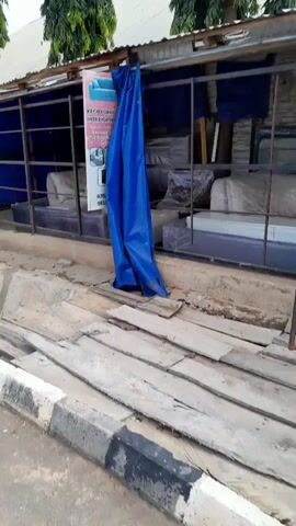 The video pans across a street-level view of a furniture shop. In the foreground, there are piles of wooden planks and a concrete curb with black and white markings. Behind the curb, a shop structure is visible, with blue tarps covering parts of the display. Shelving units hold various pieces of furniture, most of which are wrapped in plastic packaging. A sign advertising "MEGA FURNITURE INTERIOR DESIGN" is also visible. The overall impression is that of a modest, outdoor furniture store.