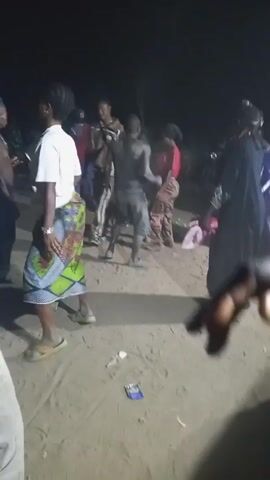 The video shows a group of people dancing outside at night. The ground is sandy, and there are cigarette packs scattered around. A person in a red baseball cap is smoking a cigarette. The dancers are wearing casual clothes and seem to be having a good time. There is one person, possibly a child, who appears to be covered in something dark. The lighting is dim, making it difficult to see the people's faces clearly.