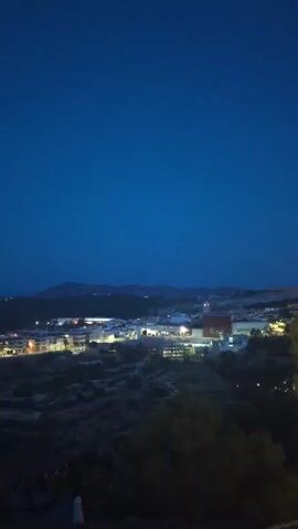 A nighttime aerial view of a town with lights illuminating buildings and streets, set against a dark, hilly landscape under a deep blue sky.