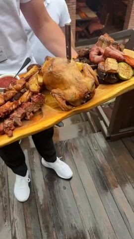 A person wearing a white t-shirt and black pants with white sneakers is holding a large wooden platter filled with food. The platter contains a whole roasted chicken, several skewers of grilled meat, roasted potatoes, sliced grilled vegetables (zucchini, eggplant, carrots), and what appears to be sausage links. There is also a small bowl of red sauce and a bowl of sliced pickles with some red onions. The person is standing on a wooden deck. In the background, there are glimpses of a restaurant interior with brick walls and wooden tables. A young boy is visible in the background. The person is holding the platter with both hands, presenting it to the viewer. A knife is sticking out of the roasted chicken.