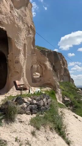 The video shows an exterior scene with cave dwellings carved into a rock face. A chair sits on a small stone enclosure in front of one of the caves. Below the cave dwellings is a hillside with some green vegetation. In the distance is a small town in a valley, and further beyond, rolling hills are visible. The sky is blue with scattered clouds. A power line is stretched across the sky above the rock face. The camera pans to the right to show more of the hillside and the town below.